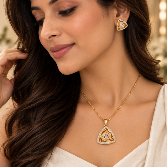 A woman wearing a gold triangular pendant necklace and matching stud earring, both featuring a geometric interwoven design with small clear stones and a central blue accent, shown in a warm, softly lit setting.