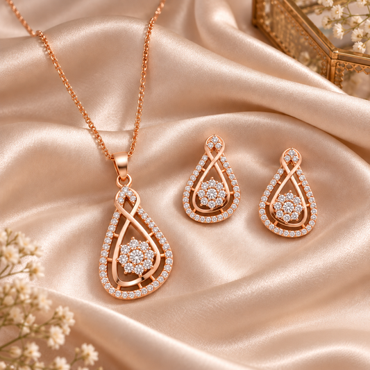 Rose gold jewelry set featuring a teardrop pendant necklace and matching drop earrings, both adorned with clear stones, displayed on draped champagne satin fabric.