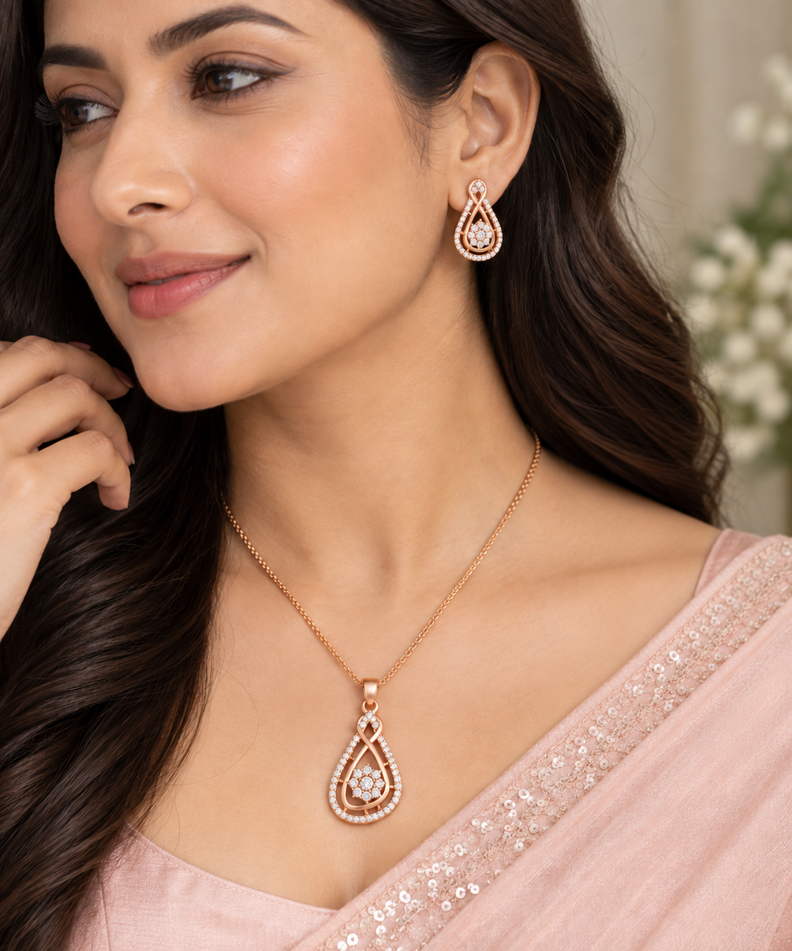A woman wearing a rose gold teardrop pendant necklace and matching drop earrings, styled with a light pink sequined sari.