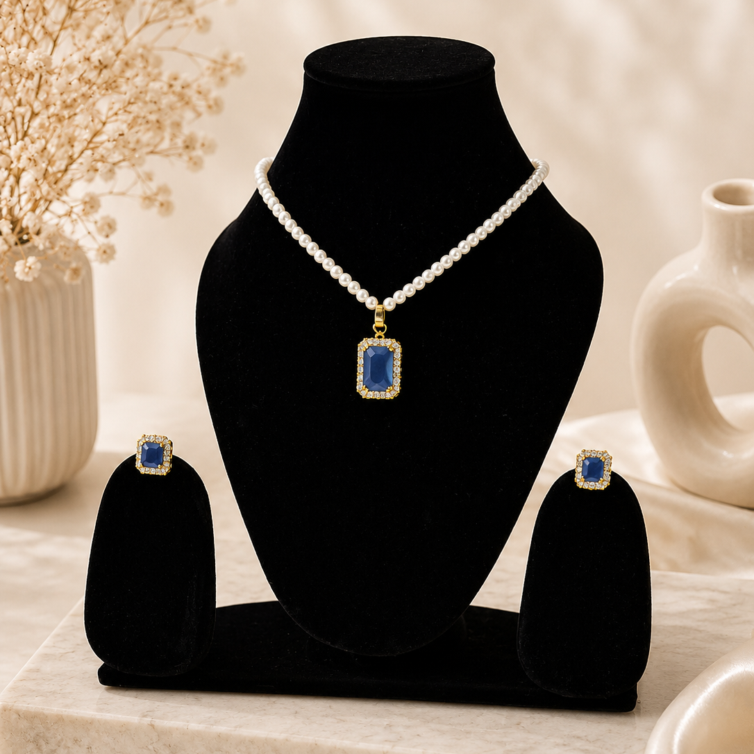 A jewelry set displayed on a black velvet bust and matching stand, featuring a pearl necklace with a rectangular blue stone pendant and matching blue stone stud earrings, styled on a marble board with dried flowers and a ceramic vase in the background.