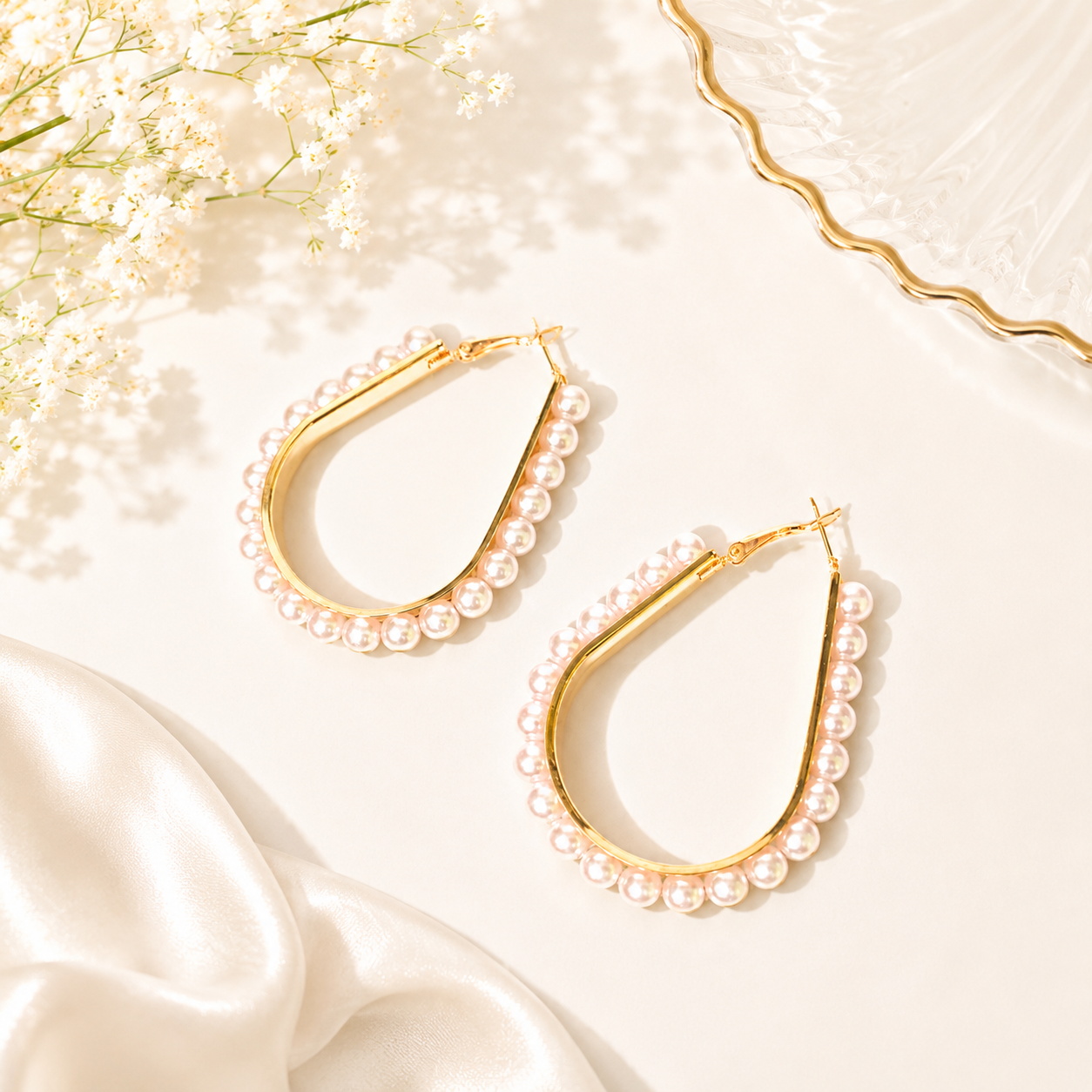 Teardrop-shaped gold hoop earrings adorned with small white pearls, displayed on a warm cream surface with soft white flowers and satin fabric accents.