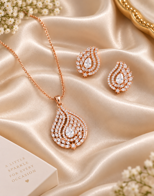 Rose gold jewelry set featuring a teardrop pendant necklace, matching stud earrings, and a small gift box, arranged on champagne satin fabric with floral accents.