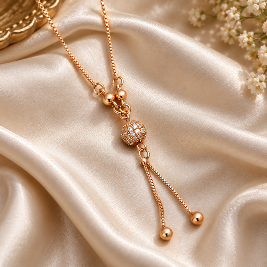 A rose gold necklace with a central pavé-set round bead and dangling beaded chains, displayed on draped champagne satin fabric with small white flowers in the corner.