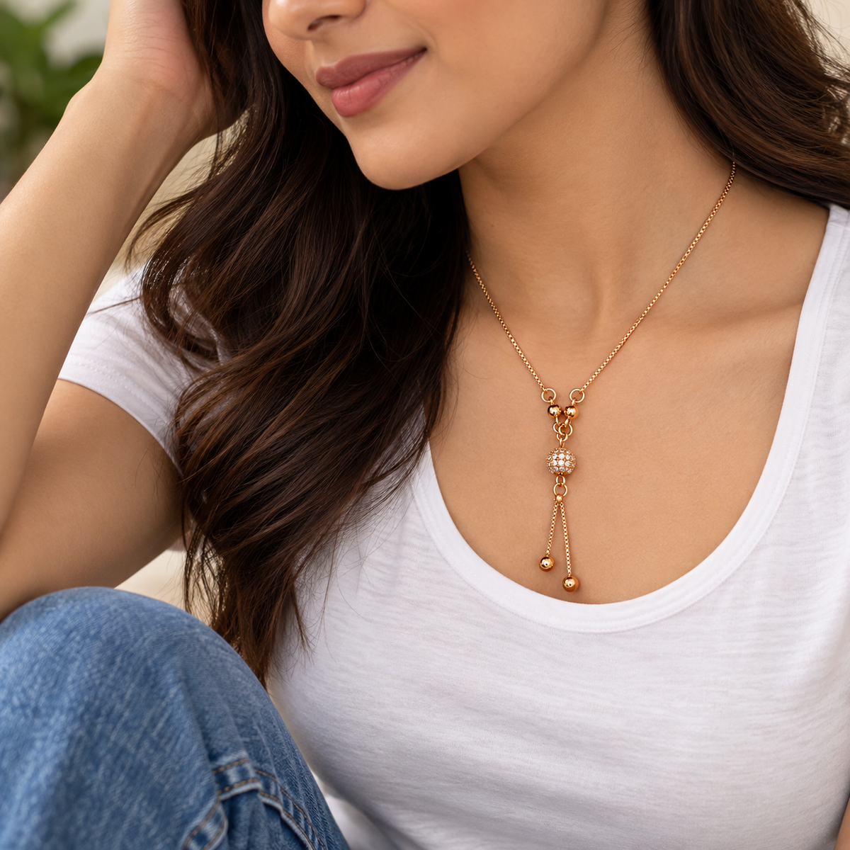 A woman wearing a delicate gold-toned necklace with a small central cluster of clear stones and two dangling bead chains, styled with a white tank top and blue jeans.