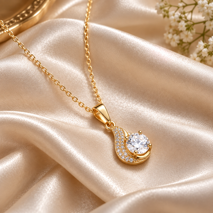 A gold pendant necklace with a crescent moon and central round clear gemstone, styled on draped champagne satin fabric with small white flowers in the corner.