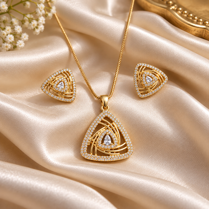 Gold-toned jewelry set featuring a triangle pendant necklace, matching triangle stud earrings, and clear gemstone accents, arranged on draped champagne satin fabric with small white flowers in the corner.