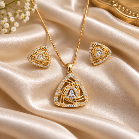 Gold-toned jewelry set featuring a triangle pendant necklace, matching triangle stud earrings, and clear gemstone accents, arranged on draped champagne satin fabric with small white flowers in the corner.