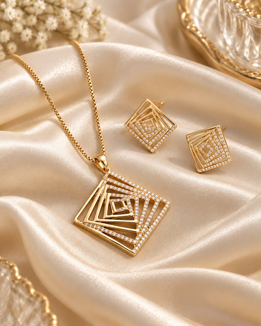 Gold jewelry set featuring a necklace and matching earrings with nested square geometric designs and clear rhinestone accents, displayed on draped champagne satin fabric.