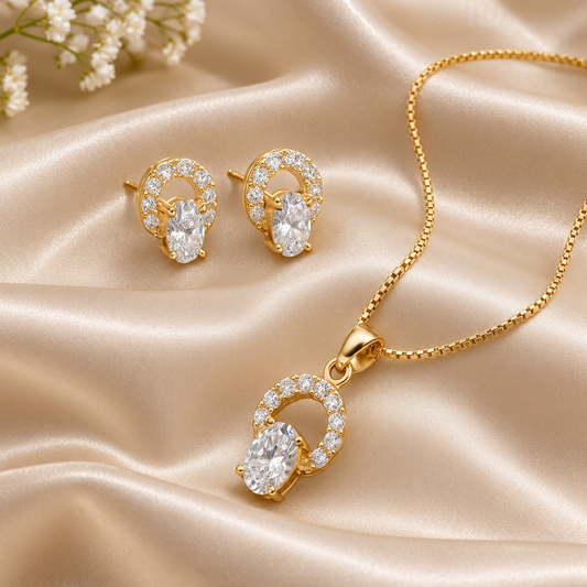 Gold jewelry set featuring a pendant necklace and matching stud earrings, both with oval clear gemstones surrounded by small sparkling stones, arranged on draped champagne satin fabric with baby's breath flowers in the corner.