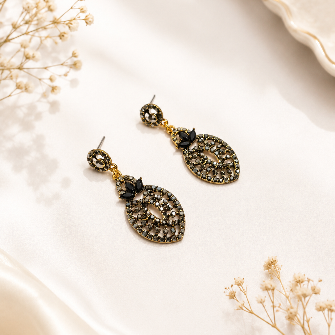 A pair of gold-toned drop earrings with oval cutout details and small black floral accents, displayed on a cream satin fabric with dried floral props.