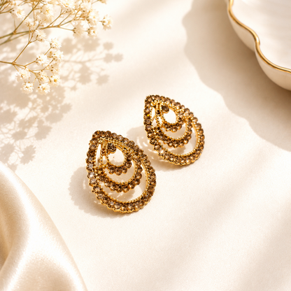 A pair of gold teardrop-shaped earrings with layered spiral patterns, displayed on a cream satin fabric with dried baby's breath flowers and a white dish in the background.