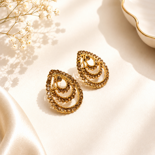 A pair of gold teardrop-shaped earrings with layered spiral patterns, displayed on a cream satin fabric with dried baby's breath flowers and a white dish in the background.