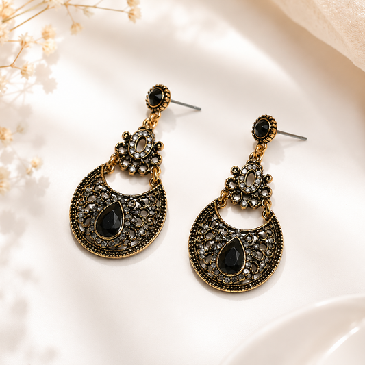 A pair of ornate gold-toned dangle earrings with black teardrop stones and silver-toned posts, displayed on a white surface with soft beige fabric and dried flowers.