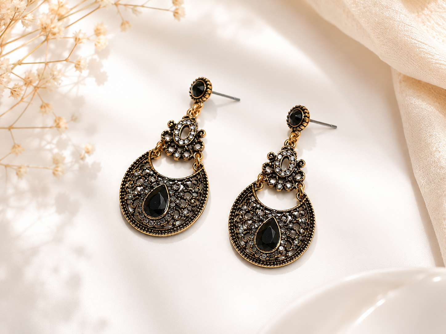 A pair of ornate gold-toned dangle earrings with black teardrop stones and silver-toned posts, displayed on a white surface with soft beige fabric and dried flowers.
