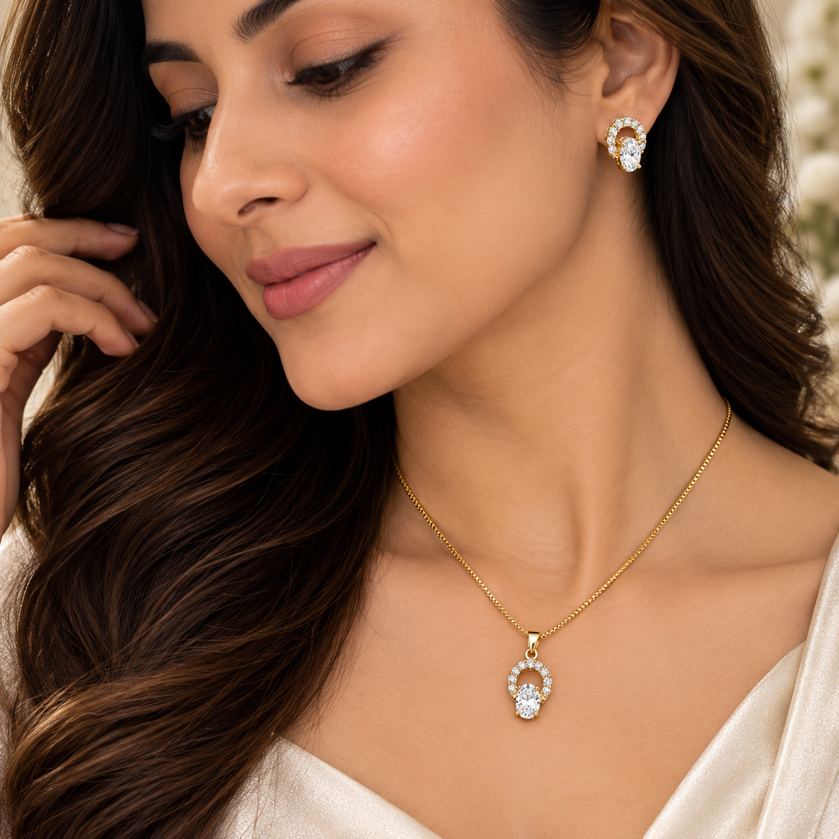 A woman wearing a delicate gold necklace with a clear teardrop-shaped gemstone pendant and a matching gold stud earring, shown in a close-up portrait.