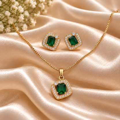 Gold jewelry set with a pendant necklace and matching stud earrings, each featuring a square emerald-cut green gemstone surrounded by small clear stones, displayed on draped champagne satin fabric with small white flowers in the corner.