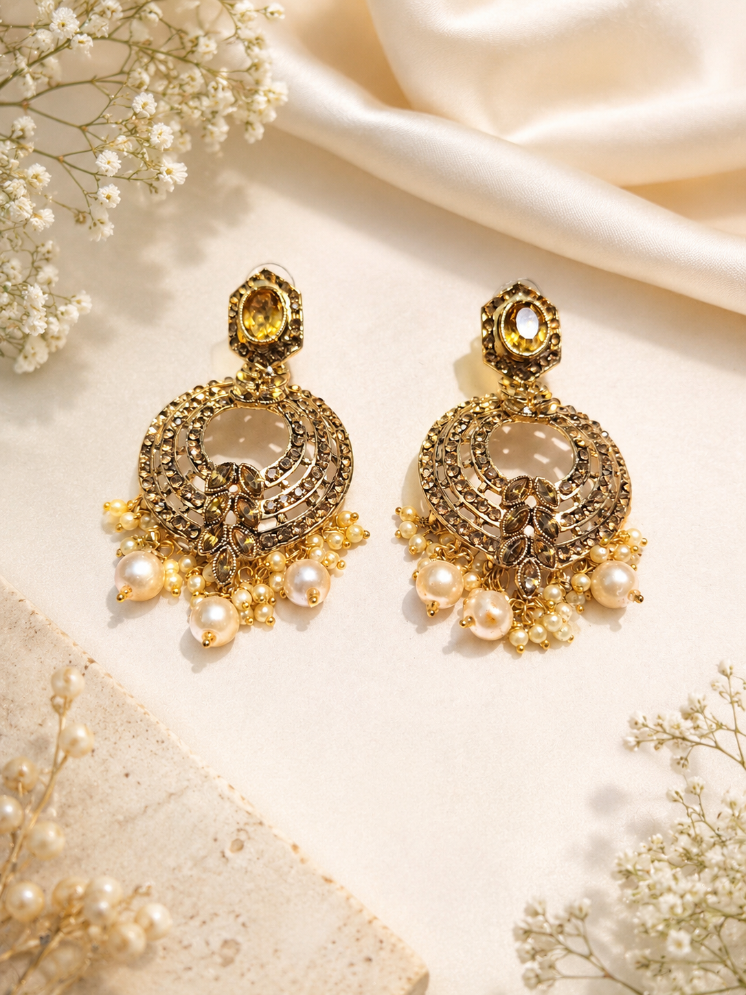 A pair of ornate gold-toned traditional Indian chandelier earrings decorated with faux pearls, crystals, and a central yellow gemstone, displayed on a cream satin fabric with dried baby's breath flowers.