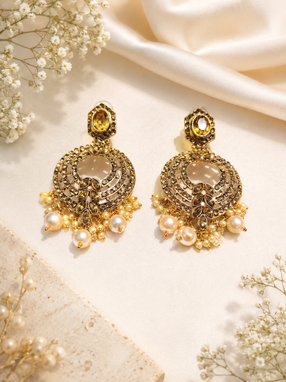 A pair of ornate gold-toned traditional Indian chandelier earrings decorated with faux pearls, crystals, and a central yellow gemstone, displayed on a cream satin fabric with dried baby's breath flowers.