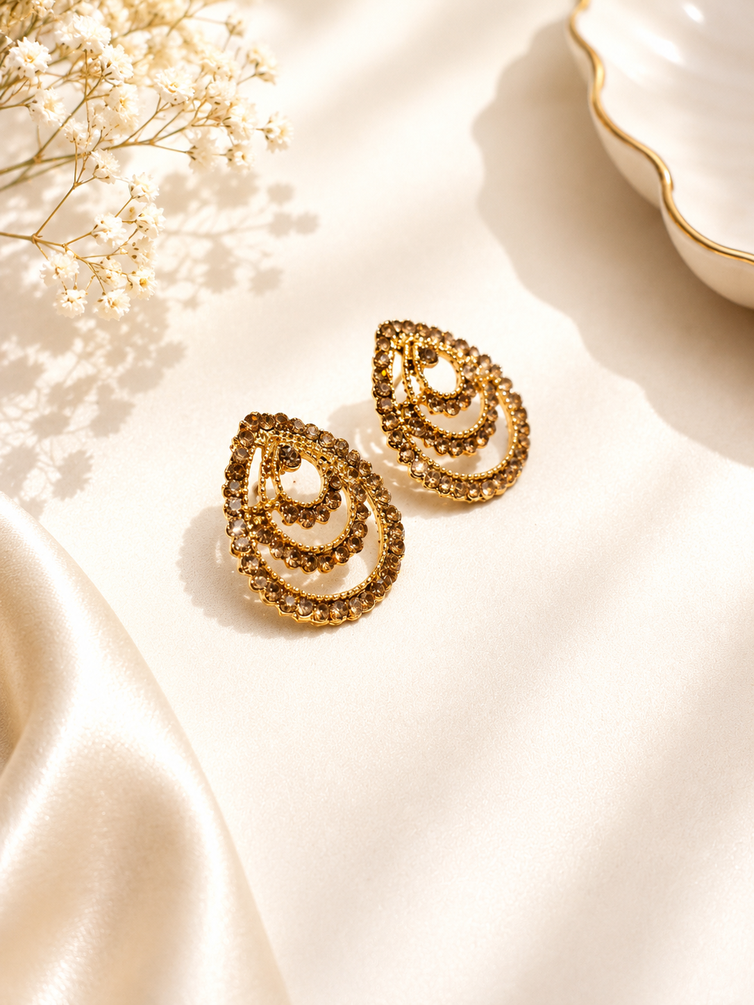 A pair of gold teardrop-shaped earrings with layered spiral patterns, displayed on a cream satin fabric with dried baby's breath flowers and a white dish in the background.