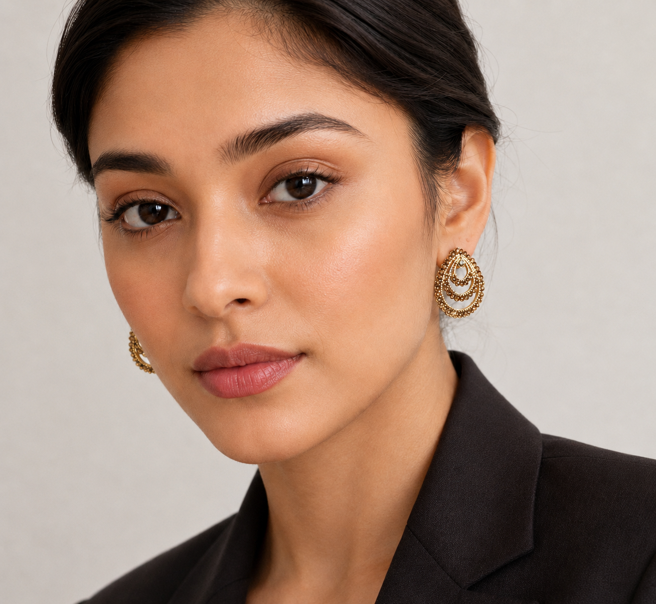 A close-up portrait of a woman wearing ornate gold crescent-shaped earrings with small clear stones, paired with a dark blazer against a soft light gray background.