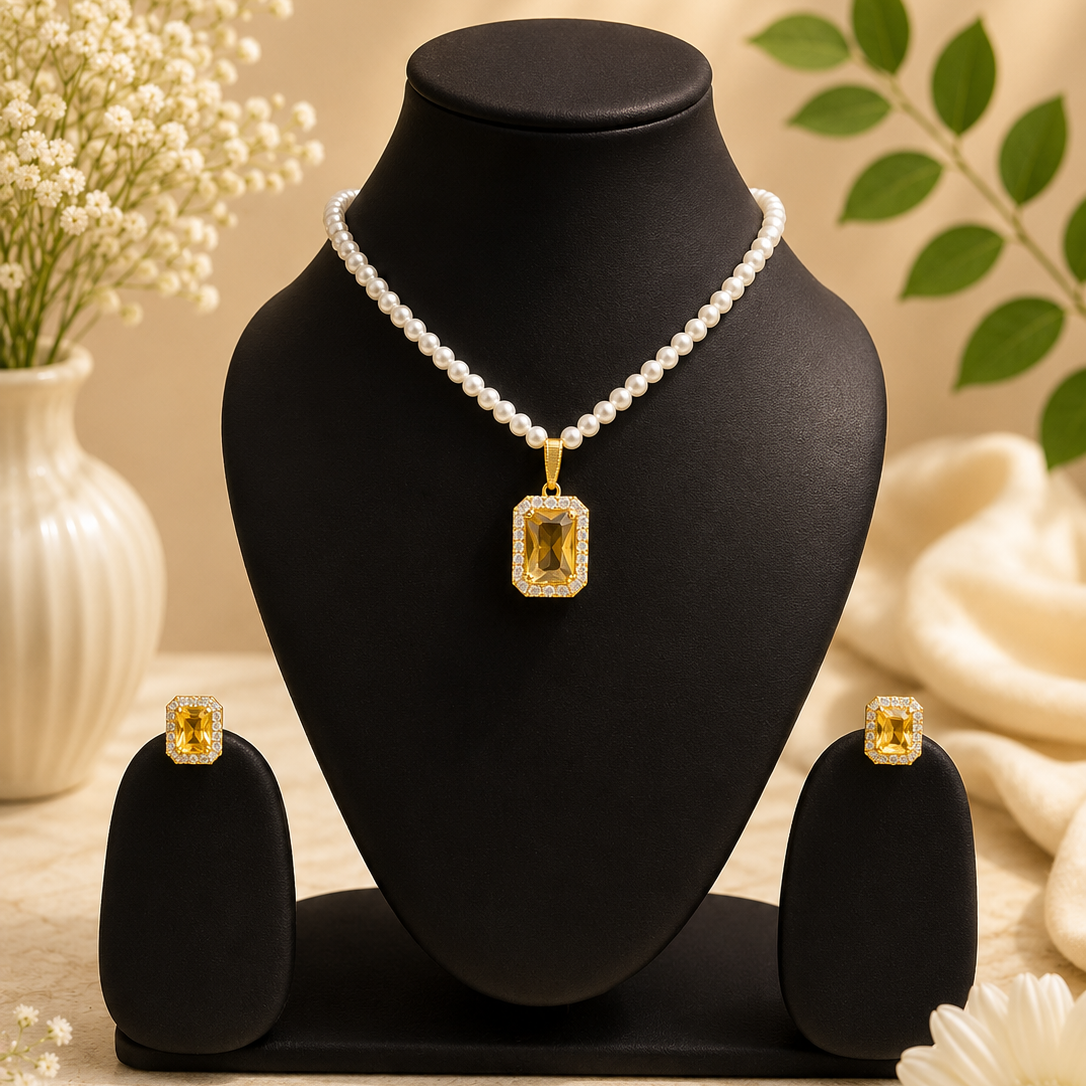 A jewelry set displayed on a black mannequin bust, featuring a pearl necklace with a rectangular yellow gemstone pendant, matching yellow gemstone stud earrings, and two black display stands. The scene is styled with white flowers and green leaves against a warm beige background.
