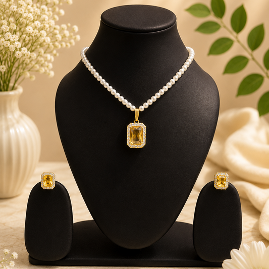A jewelry set displayed on a black mannequin bust, featuring a pearl necklace with a rectangular yellow gemstone pendant, matching yellow gemstone stud earrings, and two black display stands. The scene is styled with white flowers and green leaves against a warm beige background.