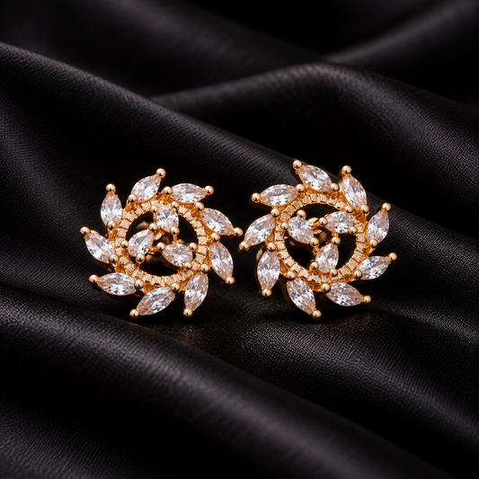 A pair of gold-toned stud earrings with marquise-cut clear stones arranged in a floral or leaf-like spiral pattern, photographed on draped black satin fabric.