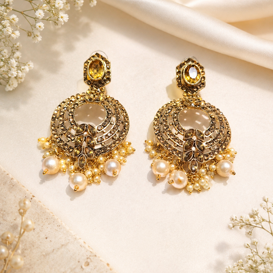 A pair of ornate gold-toned traditional Indian chandelier earrings decorated with faux pearls, crystals, and a central yellow gemstone, displayed on a cream satin fabric with dried baby's breath flowers.