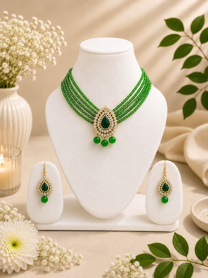 Green beaded necklace and matching drop earrings displayed on a white jewelry bust, with decorative flowers, leaves, and a candle in a warm neutral setting.