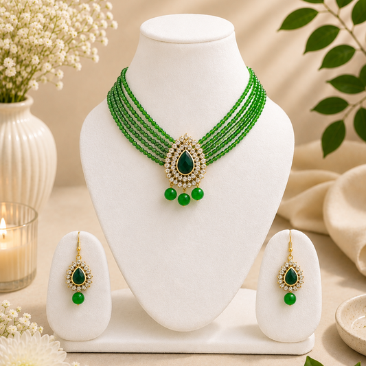 Green beaded necklace and matching drop earrings displayed on a white jewelry bust, with decorative flowers, leaves, and a candle in a warm neutral setting.