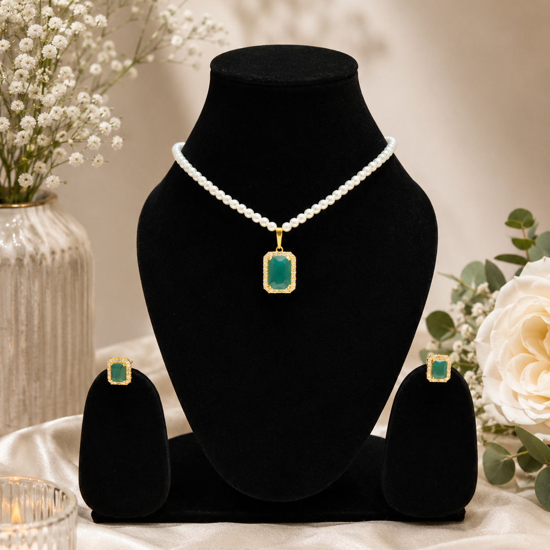 A jewelry set displayed on a black velvet mannequin bust, featuring a pearl necklace with a rectangular green stone pendant, matching green stone stud earrings, and a small matching box, styled with white flowers and a candle on a beige satin surface.