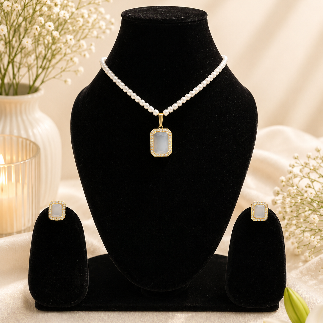 A jewelry set displayed on black velvet stands, featuring a pearl-bead necklace with a rectangular iridescent pendant, matching stud earrings, and a candle-lit floral background.