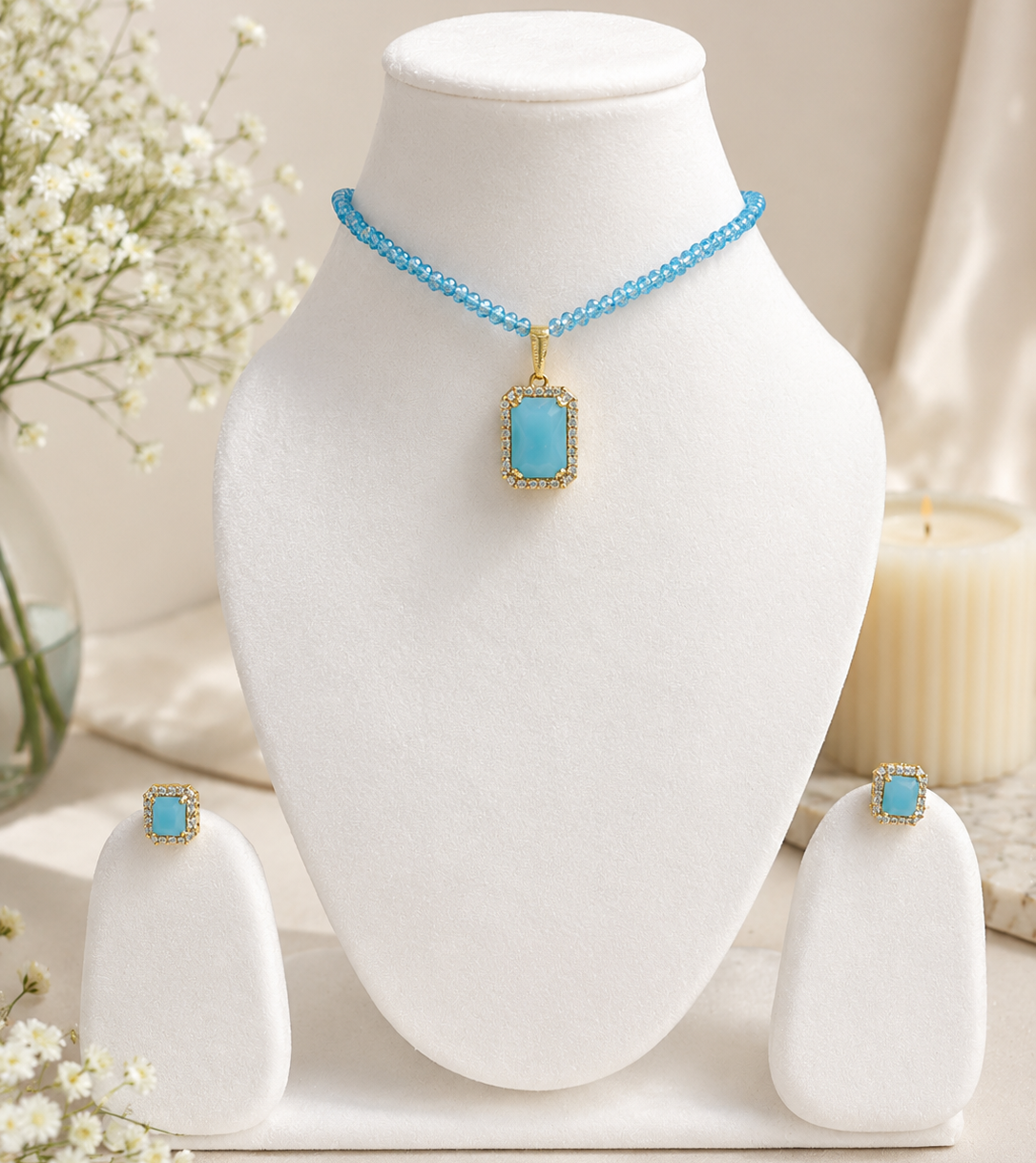 A jewelry set displayed on white mannequin stands, featuring a necklace with a rectangular light blue stone in a gold-tone frame and matching stud earrings, styled with white flowers and a candle in a soft neutral setting.