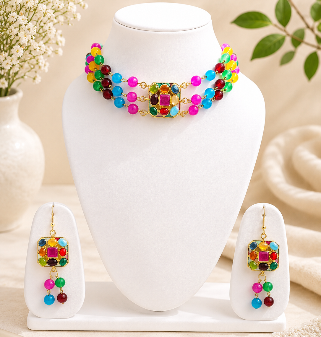 A colorful jewelry set displayed on a white mannequin bust, including a beaded necklace with a central square charm and matching drop earrings, set against a warm beige background with decorative flowers and fabric.