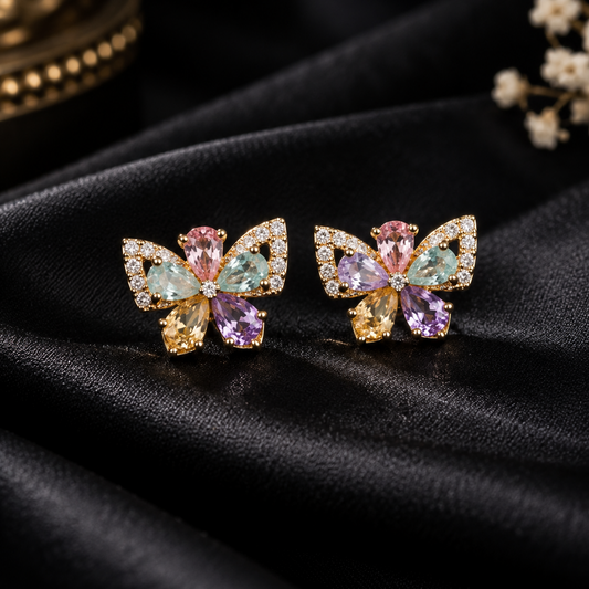 A pair of gold butterfly stud earrings with multicolored gemstones, displayed on draped black satin fabric.