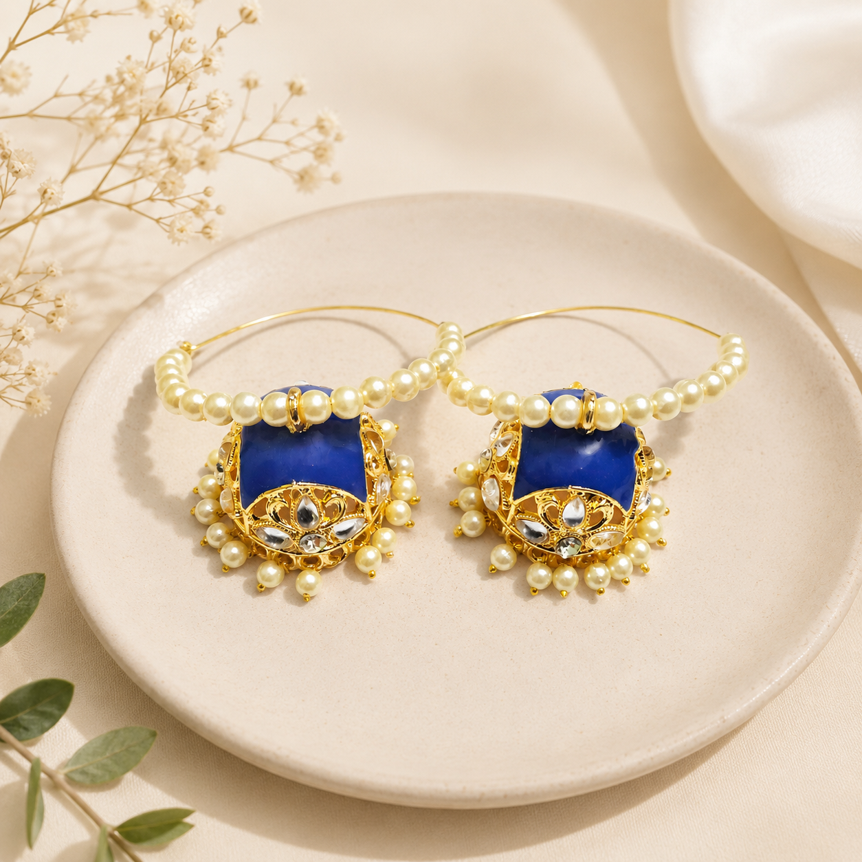 A pair of gold hoop earrings with deep blue rectangular inlays, decorated with white faux pearls and small clear stones, displayed on a round beige ceramic plate.