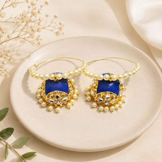 A pair of gold hoop earrings with deep blue rectangular inlays, decorated with white faux pearls and small clear stones, displayed on a round beige ceramic plate.