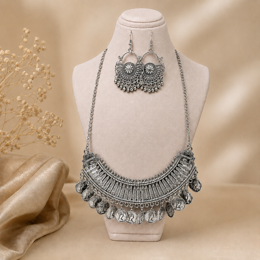 Silver-toned statement necklace and matching drop earrings displayed on a cream jewelry bust against a warm beige backdrop with decorative dried flowers and draped fabric.