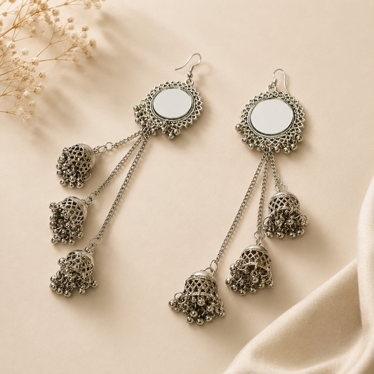 A pair of silver-toned dangle earrings with ornate filigree circular tops, small chain links, and three dangling bell-shaped charms, arranged on a warm beige fabric background with dried flowers in the corner.