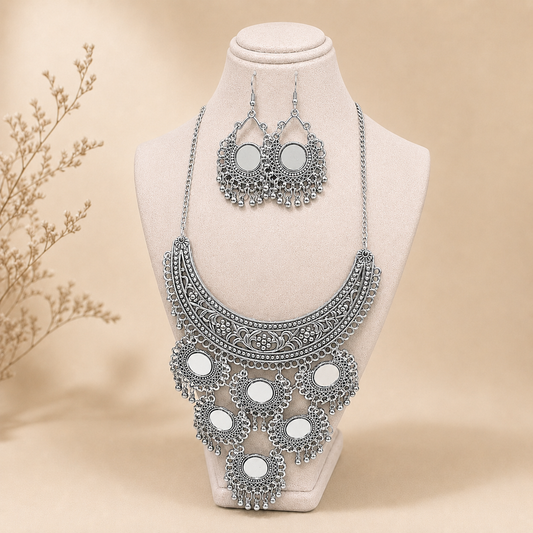 Silver-toned statement necklace and matching drop earrings displayed on a cream jewelry bust against a warm beige background with a dried floral prop at the side.