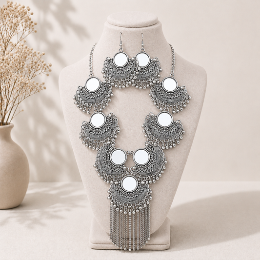 Silver-toned filigree jewelry set displayed on a cream fabric mannequin bust, including a statement necklace, matching earrings, and a choker, with white circular inlays and dangling bead chains. A beige vase with dried stems sits in the softly lit background.