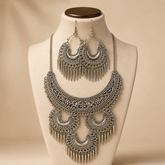 An ornate silver-toned jewelry set displayed on a cream fabric mannequin bust against a warm beige background. The set includes a large crescent-shaped statement necklace with dangling filigree and matching drop earrings.