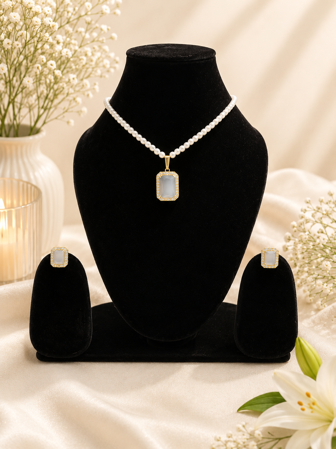 A jewelry set displayed on black velvet stands, featuring a pearl-bead necklace with a rectangular iridescent pendant, matching stud earrings, and a candle-lit floral background.