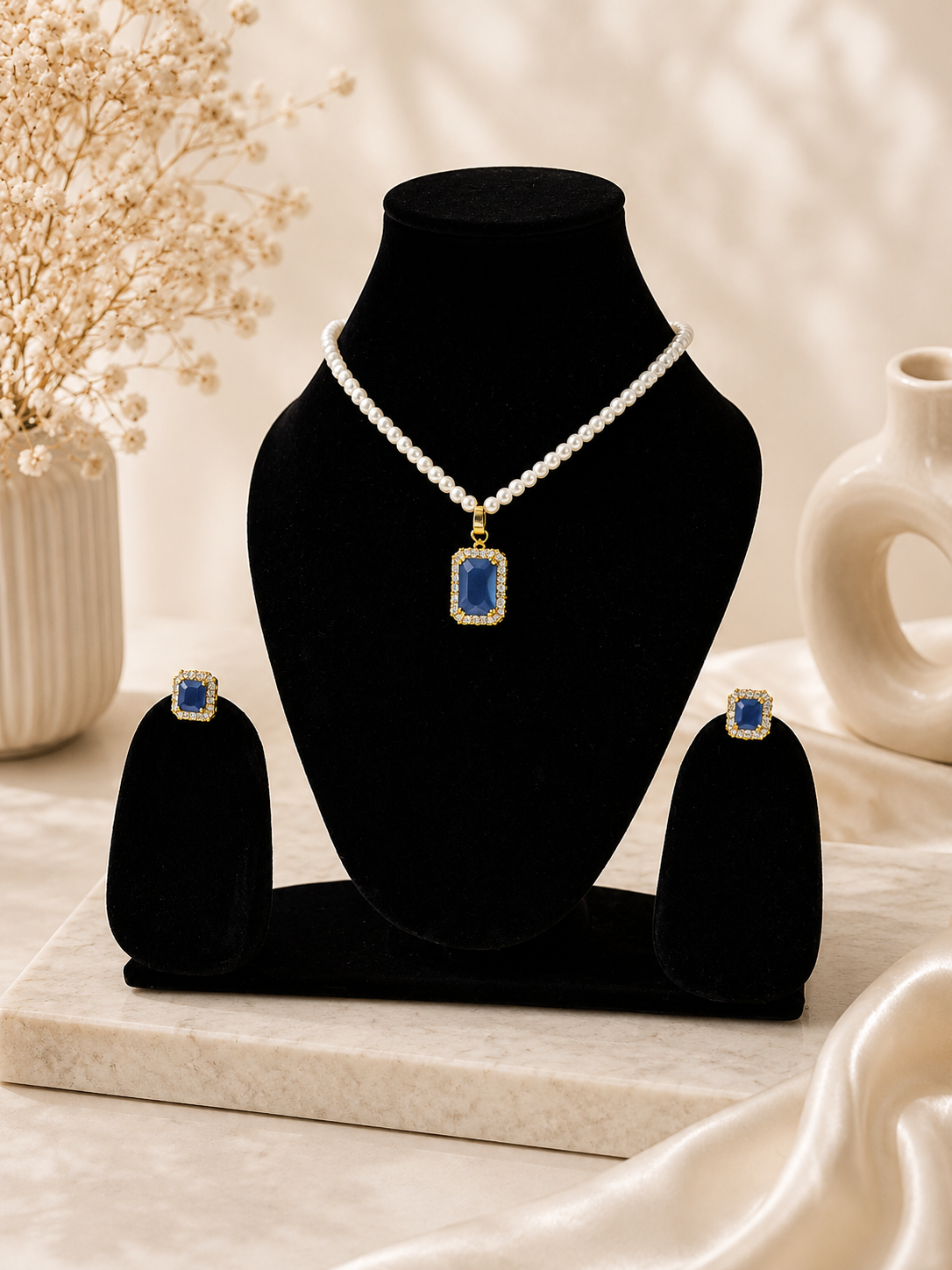 A jewelry set displayed on a black velvet bust and matching stand, featuring a pearl necklace with a rectangular blue stone pendant and matching blue stone stud earrings, styled on a marble board with dried flowers and a ceramic vase in the background.