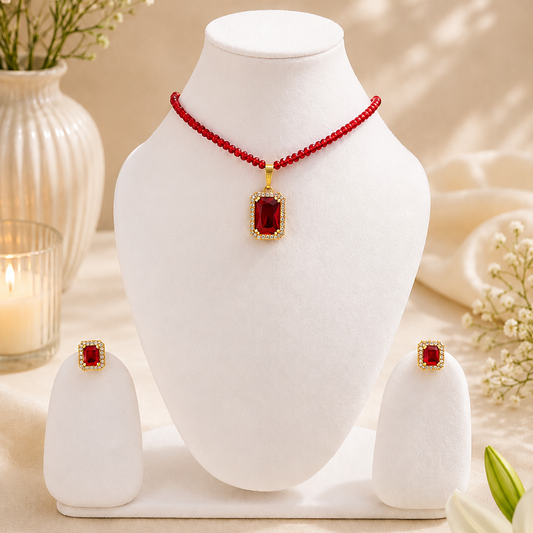 Red beaded necklace and matching stud earrings displayed on a white jewelry bust, with decorative flowers and a candle in the background.