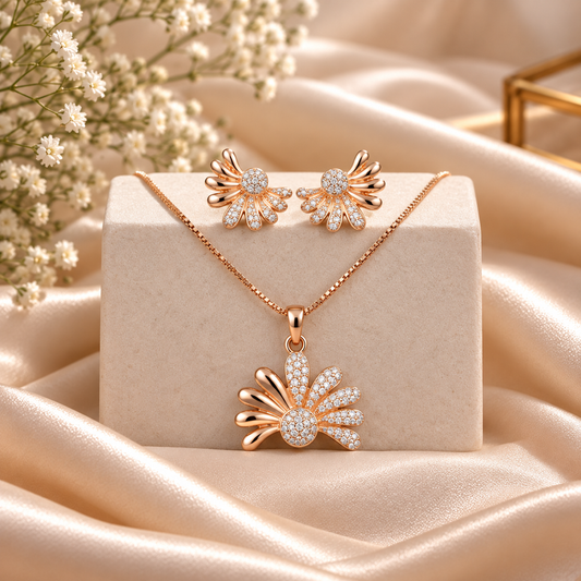 Rose gold jewelry set featuring a pendant necklace and matching stud earrings with sunburst floral designs paved with clear crystals, displayed on a cream jewelry block with champagne satin fabric and baby's breath flowers.