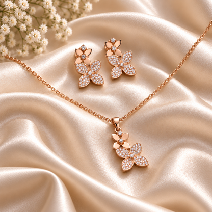 Rose gold jewelry set featuring a necklace and matching earrings with butterfly-shaped pendants and floral tops, set on draped champagne satin fabric with small white flowers in the corner.