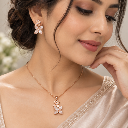 A woman wearing a rose gold flower-shaped pendant necklace and matching stud earring, both embellished with small clear stones. The jewelry is shown close-up against a soft, light background.