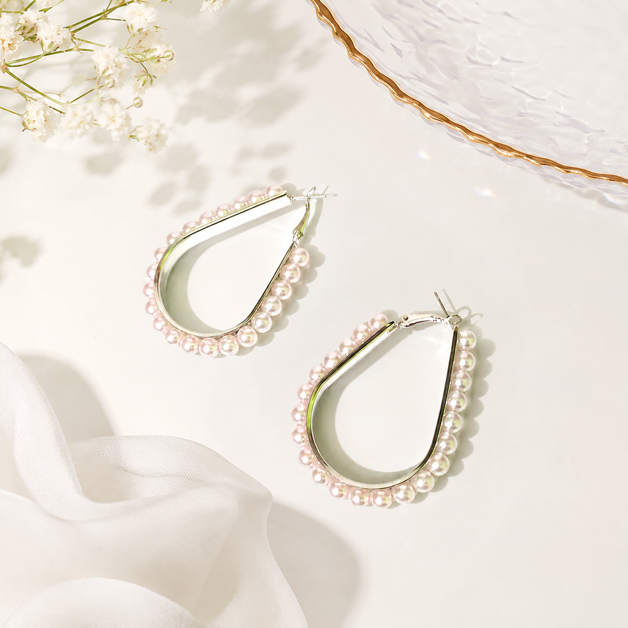 Teardrop-shaped hoop earrings with rows of small white pearls, displayed on a light cream surface with soft fabric and dried flowers as styling props.