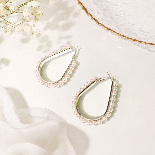 Teardrop-shaped hoop earrings with rows of small white pearls, displayed on a light cream surface with soft fabric and dried flowers as styling props.